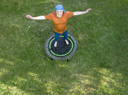 Téléchargez les photos : Senior homme en surpoids faisant de l'exercice sur un mini trampoline dans sa cour arrière, concept de remise en forme et de rebond, vue aérienne - en image libre de droit