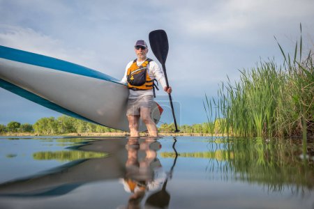 Téléchargez les photos : Senior mâle pagayeur lance un stand up paddleboard sur un lac calme au printemps, perspective grenouille à partir d'une caméra d'action au niveau de l'eau - en image libre de droit