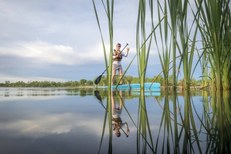 Téléchargez les photos : Senior mâle pagayeur pagaie une planche à pagaie debout sur un lac calme au début du printemps, perspective grenouille à partir d'une caméra d'action au niveau de l'eau - en image libre de droit