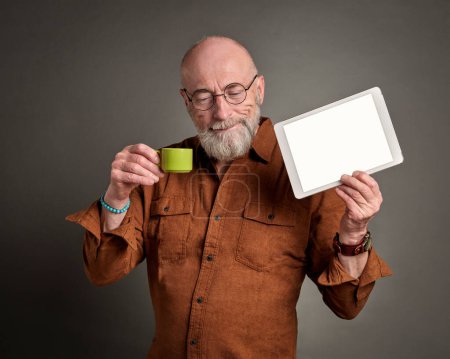 Téléchargez les photos : Souriant senior homme boit du café et montrant une tablette numérique vierge - en image libre de droit
