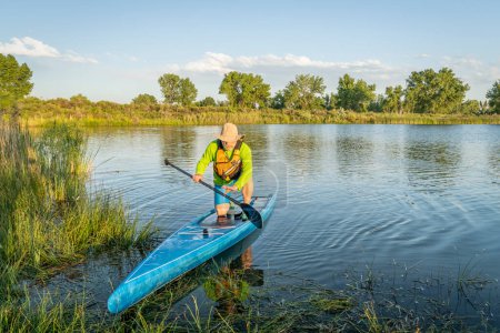Photo for Senior male stand up paddler is landing on a grassy lake shore, summer scenery in Colorado - Royalty Free Image