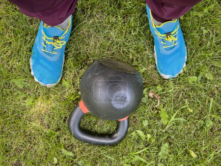 Foto de Pesadas pesas de hierro kettlebell y los pies masculinos en una hierba, concepto de fitness patio trasero - Imagen libre de derechos