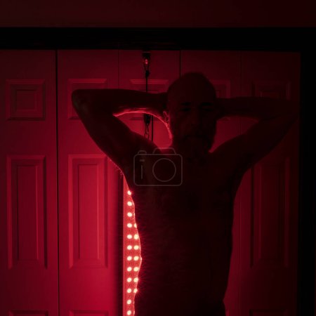 Foto de Silueta de un atlético senior, torso de hombre de pie delante del panel de terapia de luz roja, configuración del hogar para el tratamiento - Imagen libre de derechos