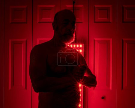 Foto de Silueta de un atlético senior, torso de hombre de pie delante del panel de terapia de luz roja, configuración del hogar para el tratamiento - Imagen libre de derechos