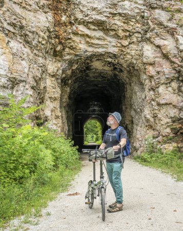 Téléchargez les photos : Homme âgé avec un vélo pliant sur Katy Trail dans un tunnel près de Rocheport, Missouri, paysages d'été. Le sentier Katy est un sentier cyclable de 237 milles converti d'un ancien chemin de fer. - en image libre de droit