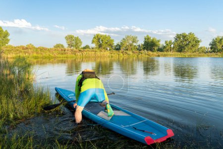 Foto de Macho stand up paddler está lanzando su paddleboard en una orilla del lago cubierta de hierba, paisaje de verano en Colorado - Imagen libre de derechos