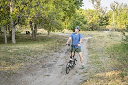 Téléchargez les photos : Athlète senior est en vélo pliant sur une route sablonneuse dans Whitetail terrain de camping dans la forêt nationale du Nebraska, paysage de la fin de l'été - en image libre de droit