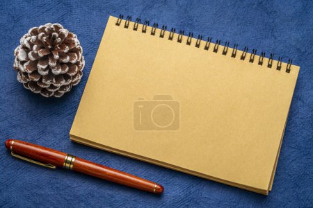 Foto de Cuaderno en espiral en blanco con un cono de pino esmerilado decorativo y una pluma de lujo sobre papel de arte - Imagen libre de derechos
