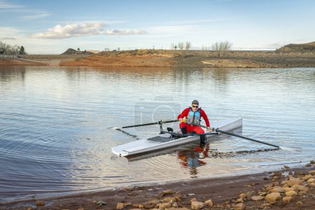 Photo for Senior male rower in a coastal rowing shell is landing on a rocky shore of Horsetooth Reservoir in fall or winter scenery in northern Colorado. - Royalty Free Image