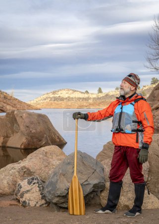 Téléchargez les photos : Pagayeur masculin senior avec une pagaie en bois sur un rivage rocheux d'un lac de montagne - réservoir Horsetooth en automne ou en hiver - en image libre de droit