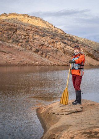 Téléchargez les photos : Pagayeur masculin senior avec une pagaie en bois sur un rivage rocheux d'un lac de montagne - réservoir Horsetooth en automne ou en hiver - en image libre de droit