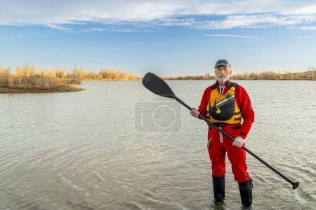Téléchargez les photos : Portrait environnemental d'un pagayeur masculin âgé en combinaison sèche et gilet de sauvetage tenant une pagaie debout sur la rive d'un lac - en image libre de droit