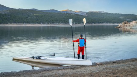 Téléchargez les photos : Un rameur mâle senior et une coquille d'aviron côtière atterrissent sur une rive rocheuse du lac Carter en automne ou en hiver dans le nord du Colorado. - en image libre de droit