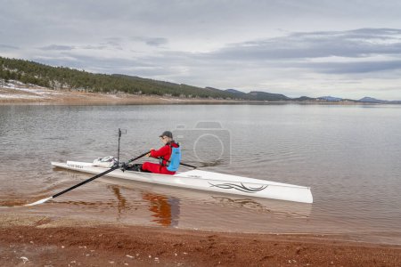 Téléchargez les photos : Loveland, CO, États-Unis - 6 février 2024 : Un rameur sénior débarque une coquille d'aviron Liteboat sur une rive du lac Carter, dans le nord du Colorado, dans un paysage hivernal. - en image libre de droit