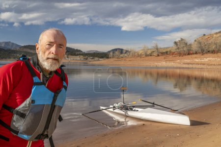 Téléchargez les photos : Portrait environnemental d'un aîné portant une combinaison sèche et un gilet de sauvetage avec une coquille d'aviron sur la rive du lac - en image libre de droit
