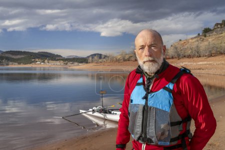 Foto de Retrato ambiental de un hombre mayor con traje seco y chaleco salvavidas con una cáscara de remo en la orilla del lago - Imagen libre de derechos