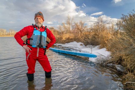 Téléchargez les photos : Pagayeur senior portant un gilet de sauvetage et une combinaison sèche est debout dans l'eau à côté de son stand up paddleboard sur le lac dans le Colorado en hiver ou au début du printemps - en image libre de droit
