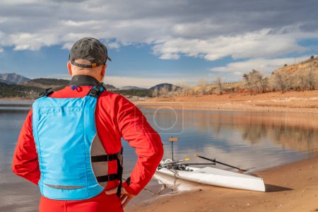 Téléchargez les photos : Senior en combinaison sèche et gilet de sauvetage avec une coquille d'aviron sur la rive du lac, l'hiver dans le nord du Colorado - en image libre de droit