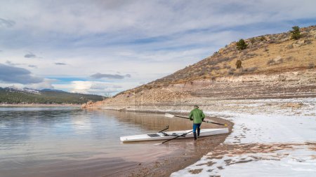 Remo macho senior con una cáscara de remo costera y remos de hacha en una orilla del lago Carter en otoño o invierno en el norte de Colorado.