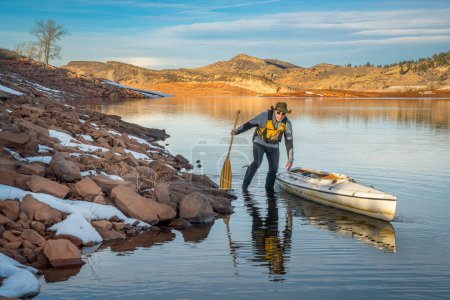 Foto de Senior male wearing life jacket is paddling expedition canoe in winter scenery of Horsetooth Reservoir in northern Colorado - Imagen libre de derechos