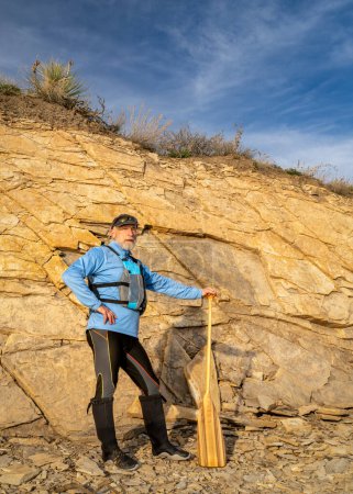 senior male paddler wearing life jacket with a wooden canoe paddle on a rocky lake shore