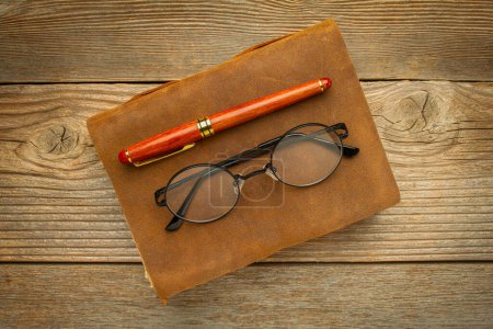 Foto de Diario de cuero antiguo con bolígrafo elegante y gafas de lectura en una mesa de madera rústica, concepto de diario - Imagen libre de derechos