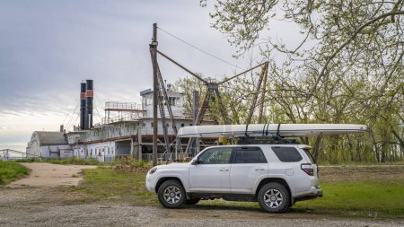 Foto de Brownville, NE, Estados Unidos - 21 de abril de 2024: Toyota 4Runner SUV con canoa de expedición en una orilla del río Missouri en paisajes de primavera con una draga histórica del río, Capitán Meriwether Lewis, en el fondo. - Imagen libre de derechos