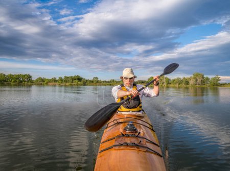 bow view of a senior male paddling a home built wooden sea kayak on a lake in Colorado