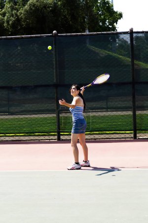 Photo for Latina Teen Woman In Jeans Skirt And Top Holding Tennis Racket On Court Serving Ball In Air - Royalty Free Image