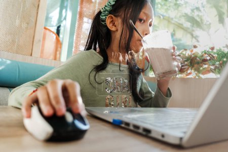 Photo for Asian teen girl drinking milkshake while using laptop in a restaurant - Royalty Free Image