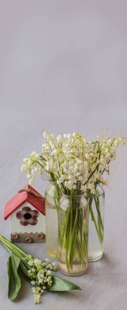 Blühende Maiglöckchen (Convallaria majalis) und dekoratives Haus auf grauem Hintergrund. Symbol des 1. Mai in Frankreich "Maiglöckchen-Tag" (Le jour du Muguet)