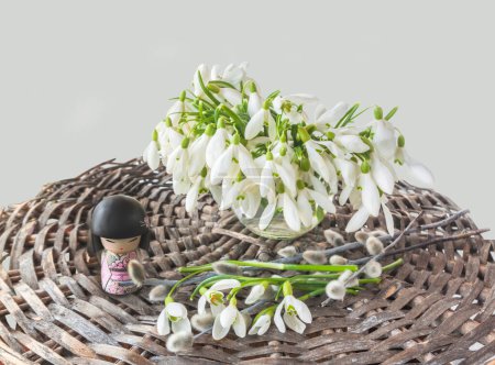 Photo for Concept of Japanese holiday of contemplating snowdrops and waiting for spring. Traditional Japanese Kokeshi doll (mass production), and snowdrops with willow branches on gray background. - Royalty Free Image