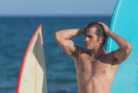 Téléchargez les photos : Un surfeur hispanique debout sur la plage en maillot de bain regardant la caméra à côté de deux planches de surf. - en image libre de droit