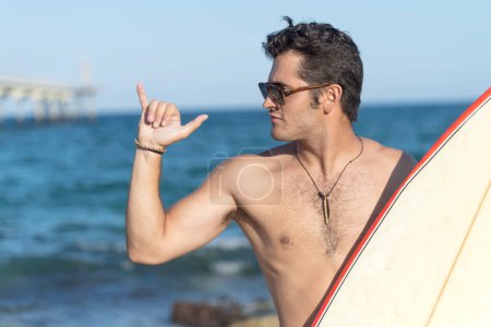 Téléchargez les photos : Surfeur hispanique garçon debout sur la plage en combinaison tenant sa planche de surf bleue et faisant le geste de surf. - en image libre de droit