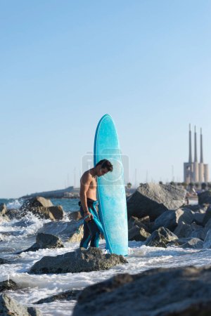 Téléchargez les photos : Surfeur hispanique garçon debout sur la plage en néoprène tenant sa planche de surf bleue au coucher du soleil - en image libre de droit