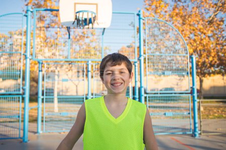 Téléchargez les photos : Portrait mi-long d'un garçon souriant dans un débardeur souriant à la caméra sur un terrain de basket urbain au crépuscule. - en image libre de droit
