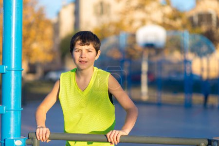 Téléchargez les photos : Portrait mi-long d'un garçon souriant dans un débardeur souriant à la caméra sur un terrain de basket urbain au crépuscule. - en image libre de droit