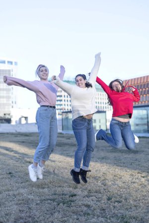 Photo for Three individuals woman, dressed in jeans and sweaters, are captured mid-jump in an outdoor field. Arms extended, they express freedom and joy. Urban buildings in the background suggest an urban park setting. - Royalty Free Image