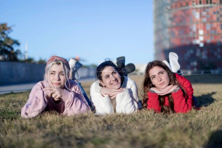 Tres mujeres jóvenes tumbadas boca abajo sobre la hierba, posando juguetonamente con las manos bajo la barbilla, con un cielo despejado y un fondo urbano