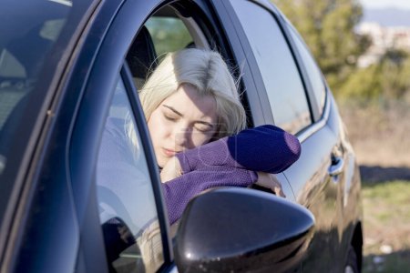 Blonde Frau, die Kopf auf Autoscheibe ruht, Augen geschlossen, in einem Moment der Entspannung oder Müdigkeit, trägt einen lila Pullover, heitere Atmosphäre