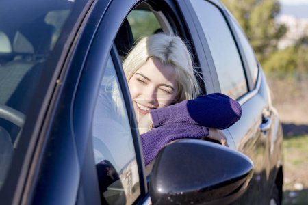 Blonde Frau, die Kopf auf Autoscheibe ruht, Augen geschlossen, in einem Moment der Entspannung oder Müdigkeit, trägt einen lila Pullover, heitere Atmosphäre