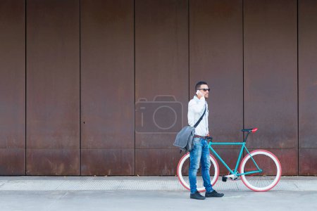 Stylischer Mann mit Sonnenbrille und Umhängetasche steht neben einem roten Fahrrad und telefoniert vor einer verrosteten Metallwand