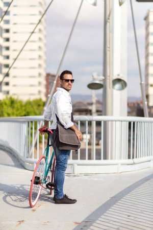 Casual man in sunglasses stands with his fixed-gear bike on a modern bridge, cityscape behind him.