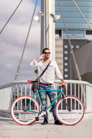 Fashionable man in white shirt and sunglasses stands with a colorful bicycle on an urban bridge, high-rise buildings in the background