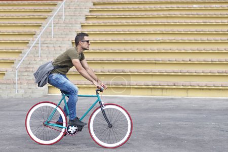 Foto de Hombre enfocado en atuendo casual monta una bicicleta de engranaje fijo en un entorno urbano, con una bolsa de mensajero sobre su hombro, retratando el transporte activo y ecológico - Imagen libre de derechos