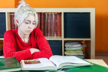 Photo for Young woman in red studying hard at a library desk, looking exhausted with books and shelves in the background, displaying academic fatigue. - Royalty Free Image