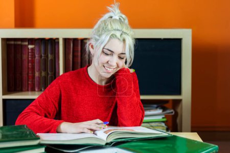 Junge Frau mit blonden Haaren, lächelnd, während sie Notizen schreibt. Im roten Pullover, am Büchertisch mit Büchern lernen, inmitten des akademischen Stresses fröhliche Stimmung verbreiten.