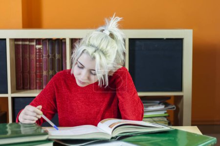 Junge Frau mit blonden Haaren, lächelnd, während sie Notizen schreibt. Im roten Pullover, am Büchertisch mit Büchern lernen, inmitten des akademischen Stresses fröhliche Stimmung verbreiten.