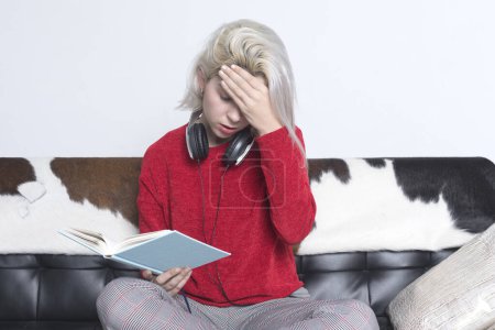 Young woman with blonde hair, in a red sweater and plaid pants, sits on a couch with a book and headphones, holding her head in a thoughtful and stressed pose.