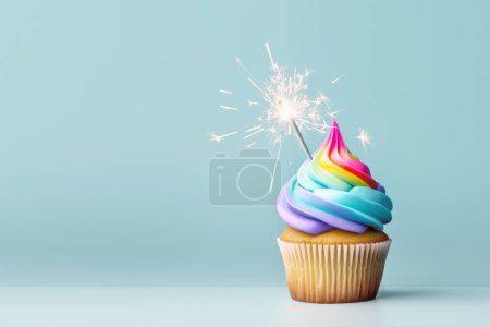 Birthday cupcake with colorful rainbow frosting and celebration sparkler for a birthday party, plain blue background with copy space to side